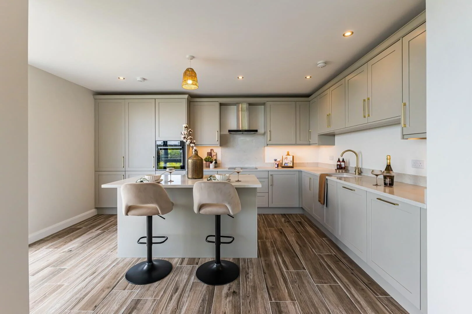 Modern kitchen with light gray cabinets, white countertops, wood flooring, center island with two beige bar stools, and decorative items including a large vase with flowers.