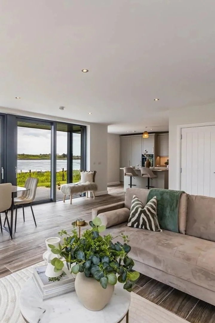 Living room with beige sofa, green and zebra-patterned pillows, round coffee table with potted plants, and a view of a lake through large sliding glass doors, with kitchen in the background.