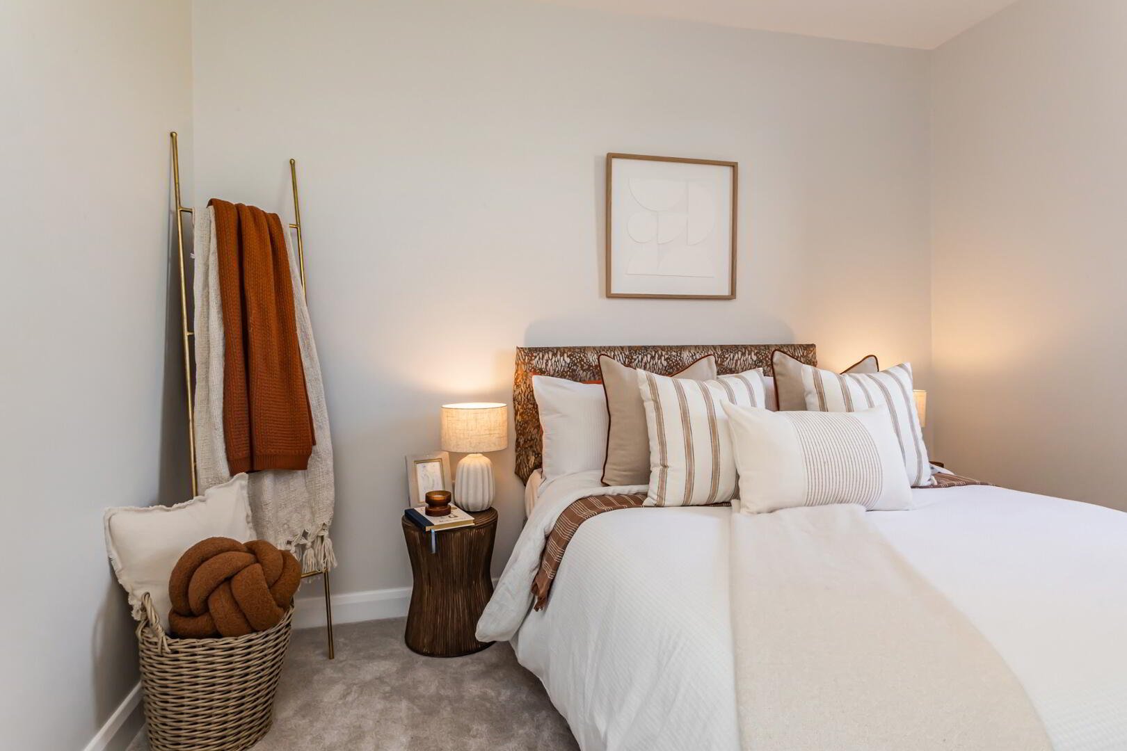 A neatly made bed with white bedding and multiple pillows in neutral tones, a wooden nightstand with a lamp, framed artwork above the bed, a gold towel rack with orange and cream blankets, and a laundry basket with a large knotted orange blanket insi