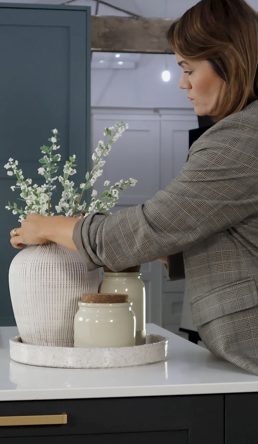 A woman arranges white flowers in a textured, beige ceramic vase on a kitchen counter, surrounded by smaller beige jars, with a blue cabinet and exposed wooden beam in the background.