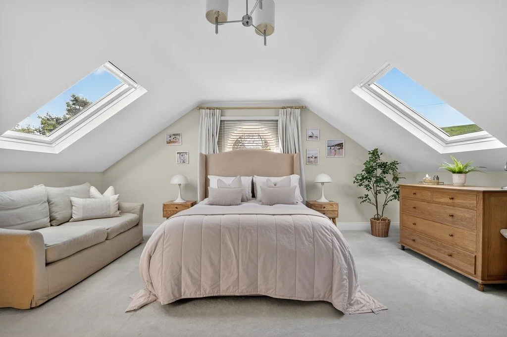 A cozy bedroom with a bed, beige headboard, and white bedding, flanked by nightstands and lamps, with a beige sofa, wooden dresser, potted plant, and skylight windows showing blue sky and trees.