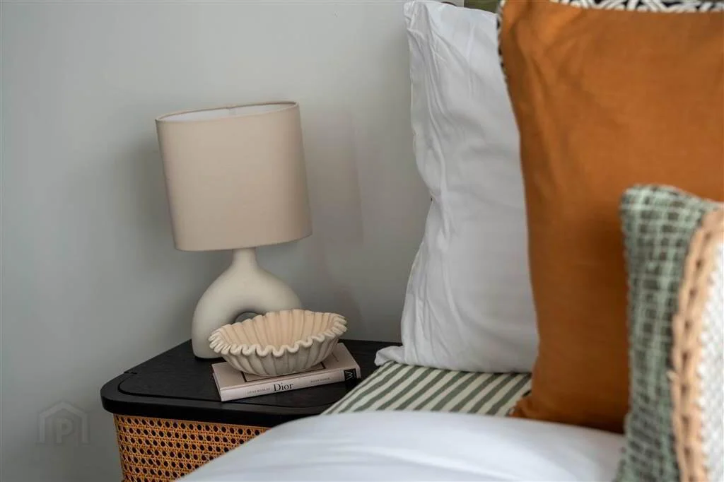 Close-up of a bedroom nightstand with a modern lamp, a decorative bowl, and a book, next to a bed with white and orange pillows.