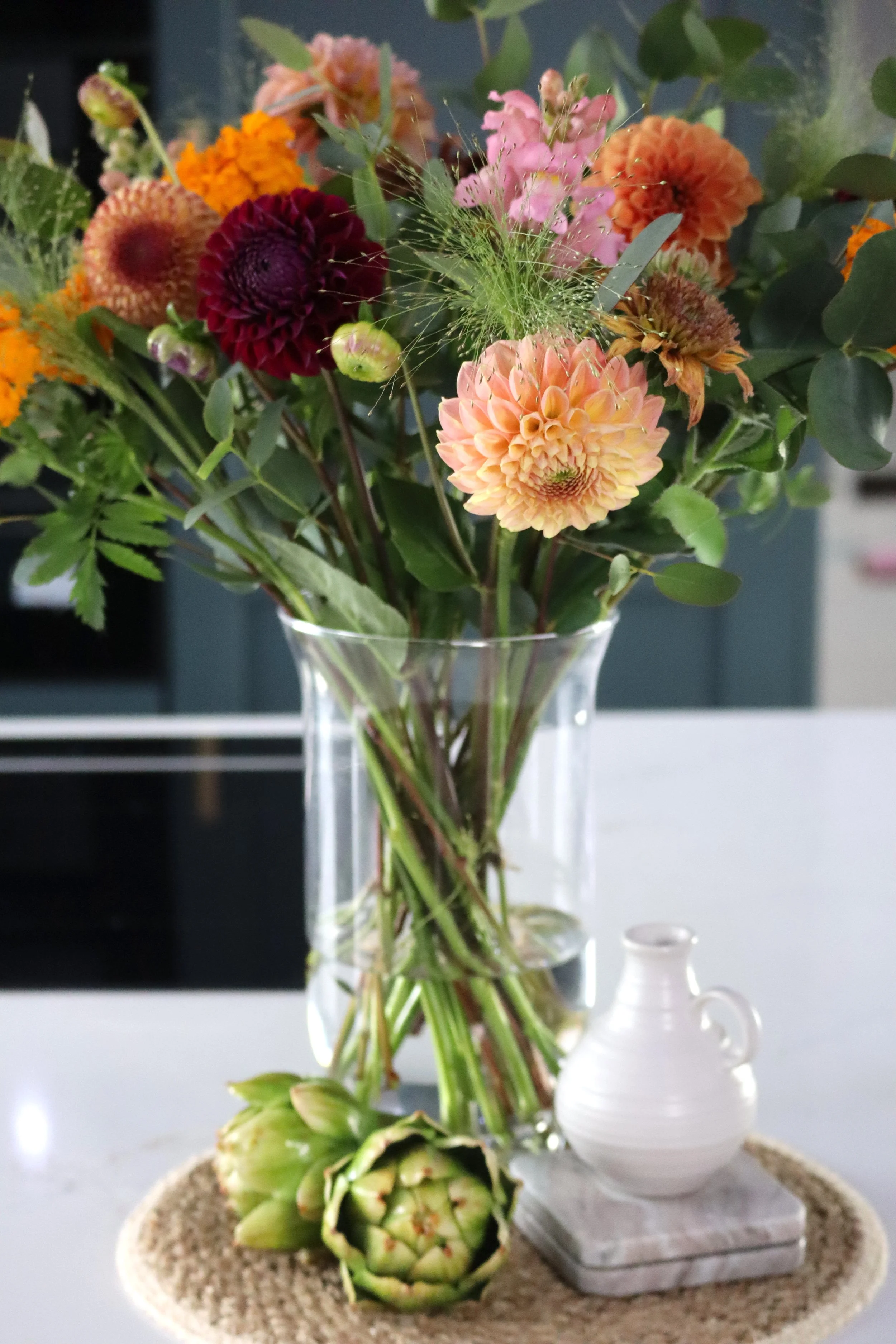 A clear glass vase filled with colorful flowers, including dahlias and other blooms, sits on a woven coaster on a white surface, with a small white ceramic jug and artichoke on the side.