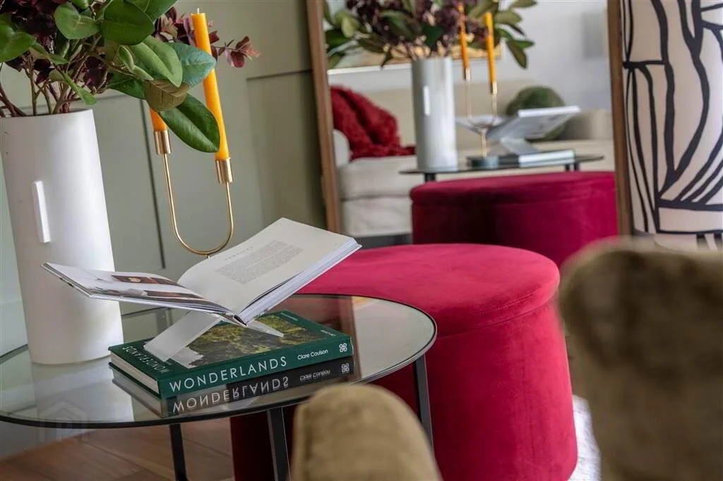 A living room with a small glass coffee table holding a book titled "Wonderlands" and an open magazine, with a tall white vase containing green leaves and purple flowers, and a yellow candle holder with candles. In the background, there are pink upho
