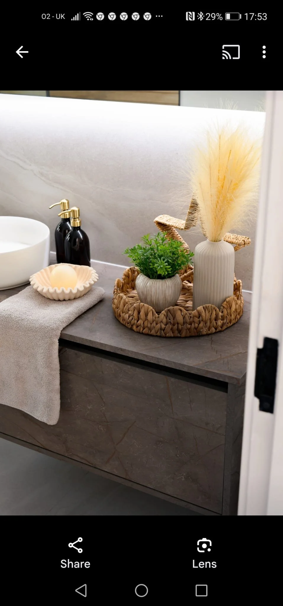 Decorative bathroom countertop featuring a white vase with pampas grass, a small plant in a striped pot, and two black soap dispensers with gold pumps, with a folded towel and a candle nearby.