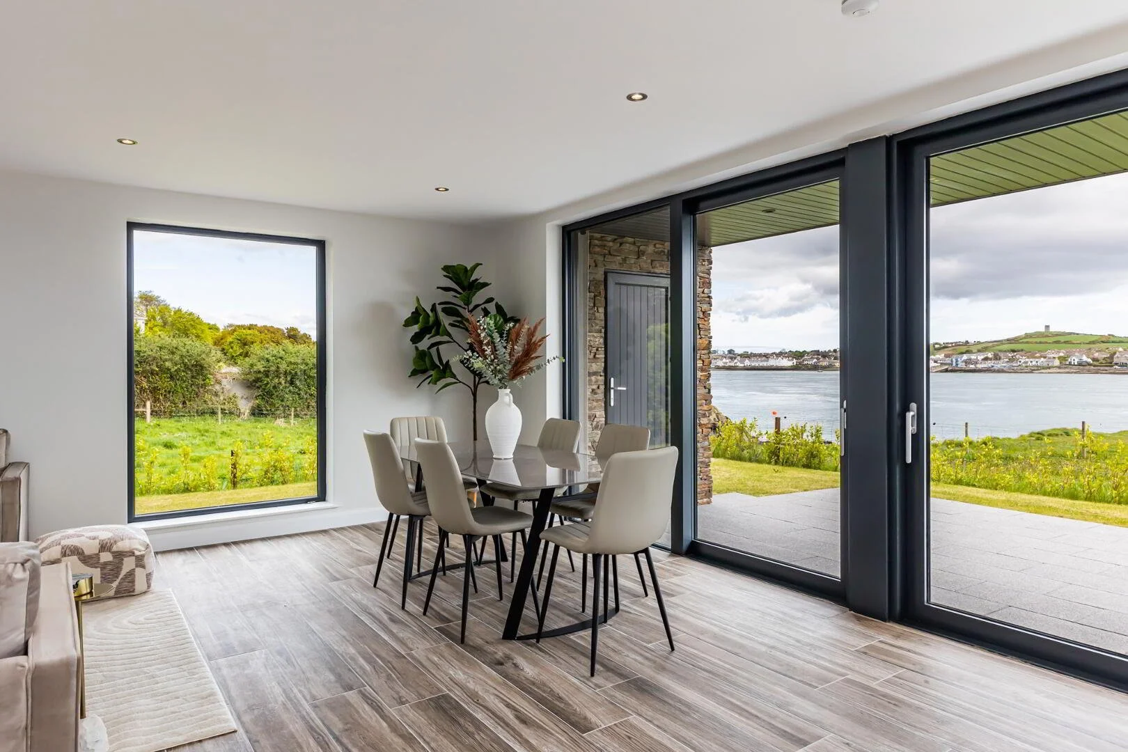 Modern dining room with large glass sliding doors overlooking a river and green landscape, beige chairs around a black dining table, and a large potted plant inside.
