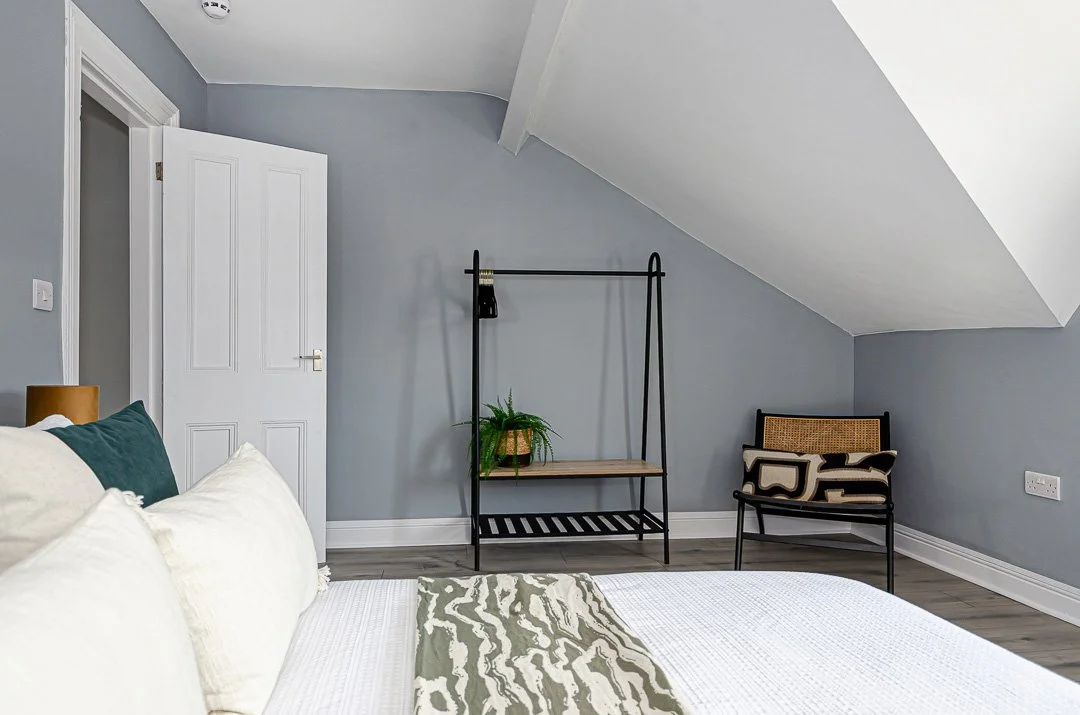Bedroom with gray walls, white door, bed with white and green pillows, a decorative throw, a black metal clothing rack with a plant on it, and a black chair with a patterned pillow.