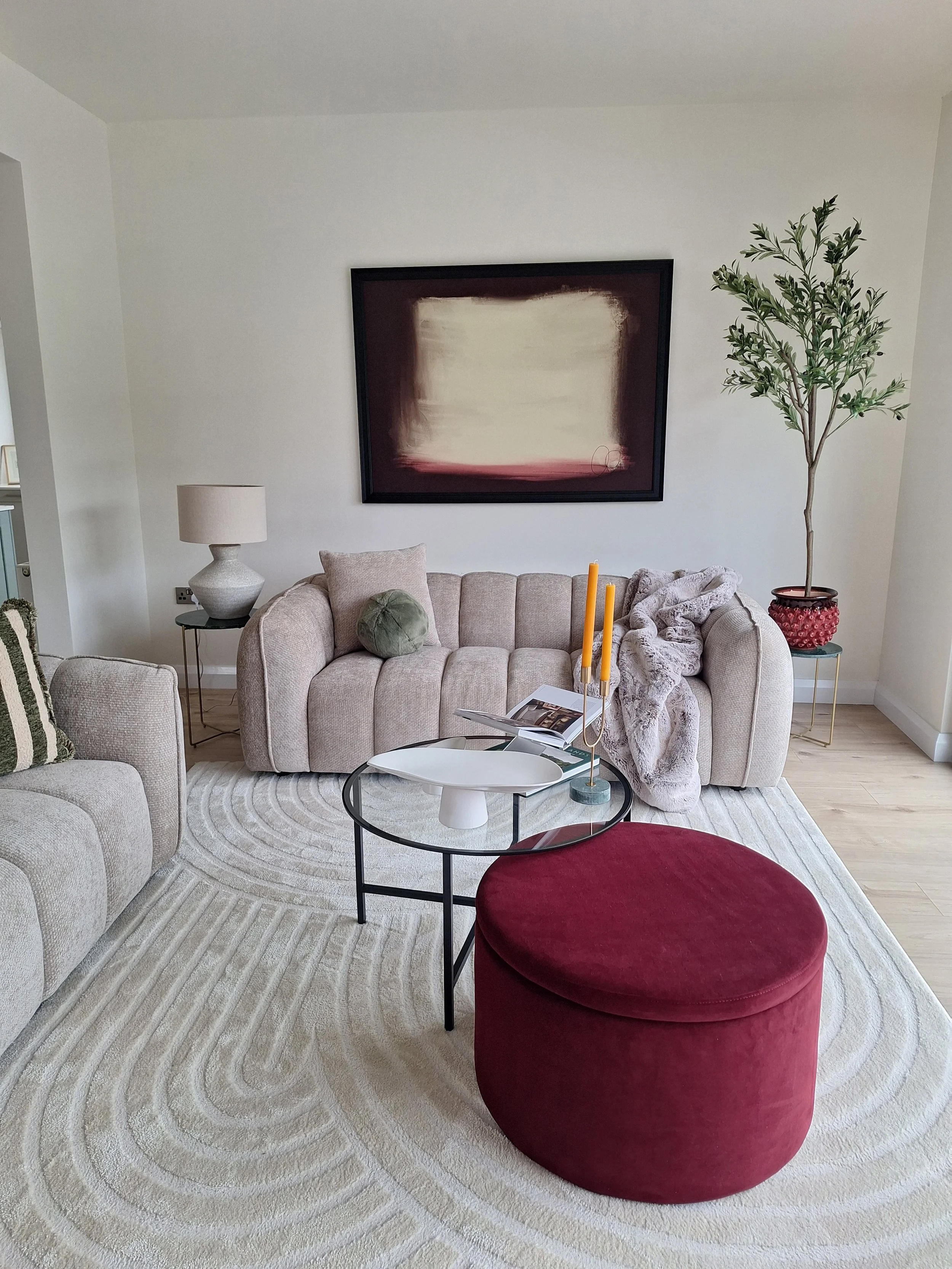 Living room with a beige sofa, a black-framed abstract painting on the wall, a glass coffee table holding a white decorative item, a book, and yellow candles, a round red velvet ottoman, a beige armchair with a striped pillow, a side table with a lam