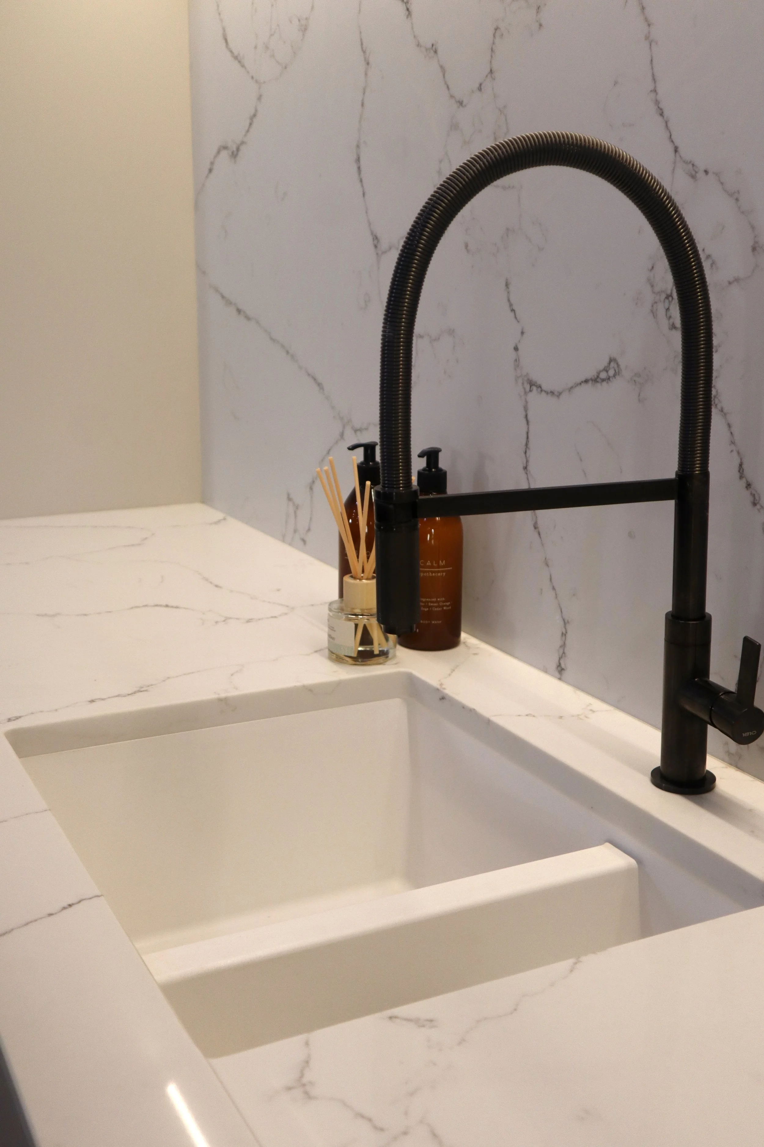Modern kitchen sink with black faucet, on white marble countertop, beige wall, and marble backsplash, with a candle and a soap dispenser beside the sink.
