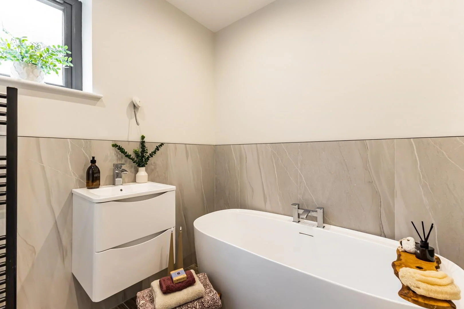 Modern bathroom with a white bathtub, a small white sink with a cabinet, a window with a plant, and decorated with towels and natural elements.