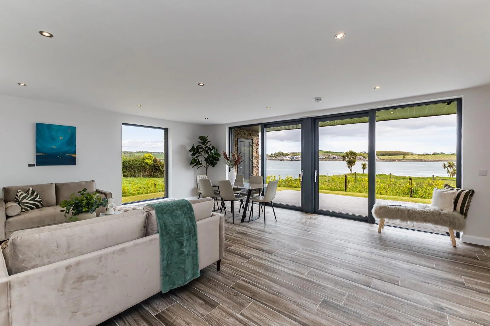 Living room with large glass sliding doors overlooking a lake and greenery, beige sofas, a small bench with a furry cushion, a dining table with chairs, and decor including a painting and a potted plant.