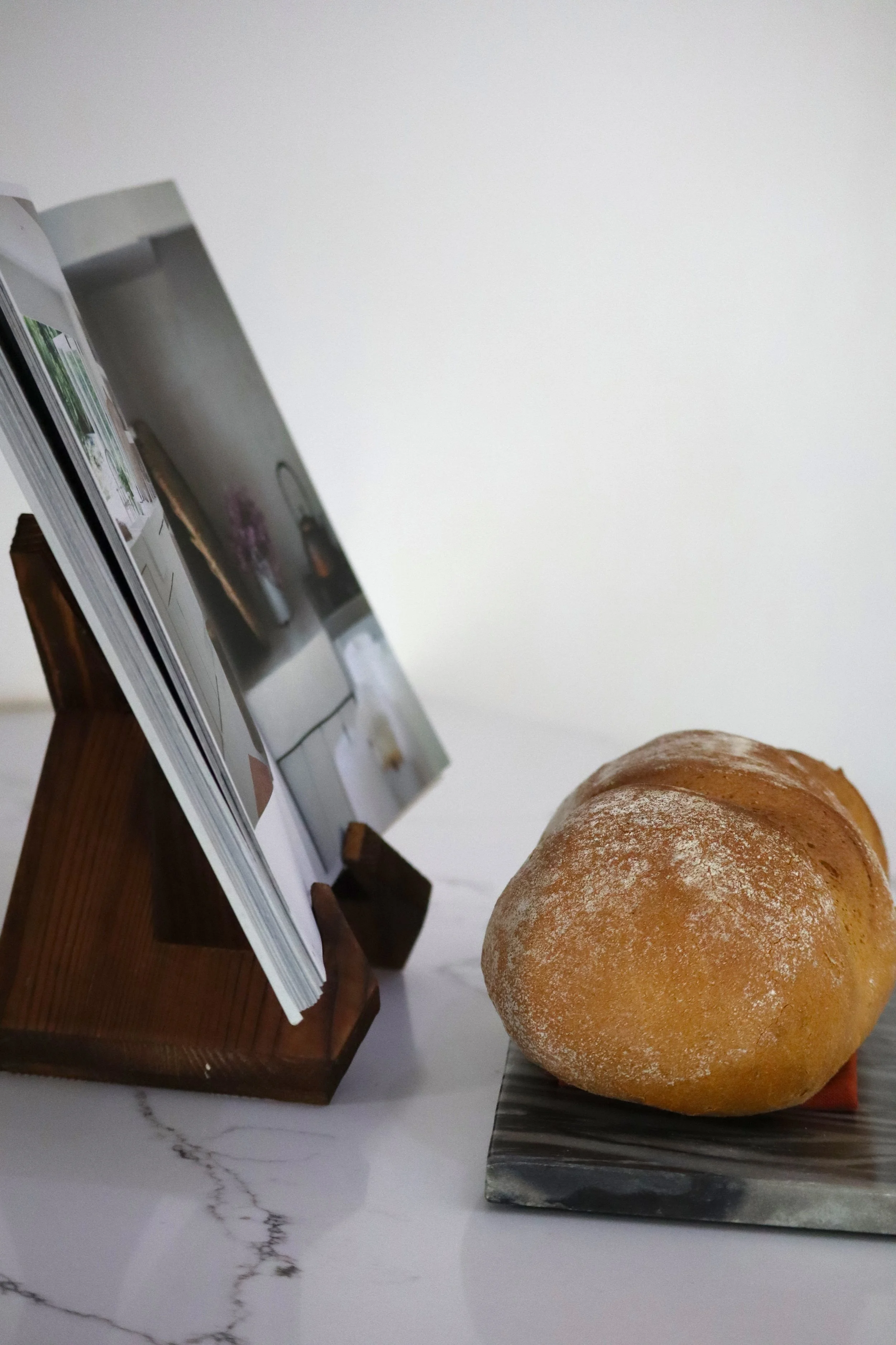 A loaf of bread with a dusting of flour on top, placed on a small dark rectangular plate, next to a wooden recipe or photo stand with a book or magazine open on it, on a white marble surface.