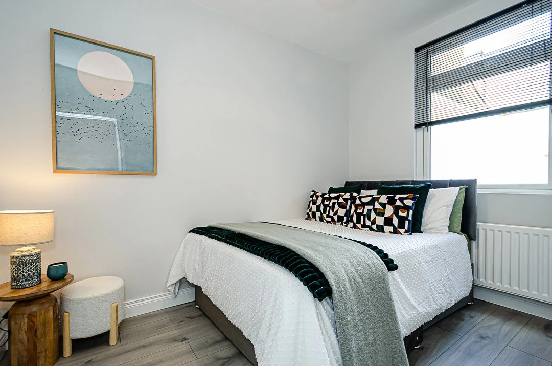 A bedroom with a bed, decorative pillows, and a window with blinds. There is a framed artwork on the wall and a nightstand with a lamp and a small decorative object.