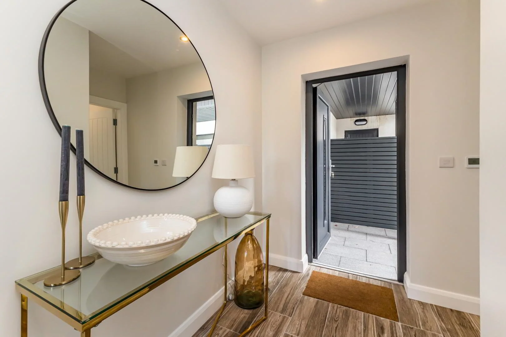 Entryway with a large round mirror on the wall, a glass console table with gold accents, a white decorative bowl, a white lamp, and a large greenish-brown vase. Open door leads to an outdoor area with a dark door and black siding.