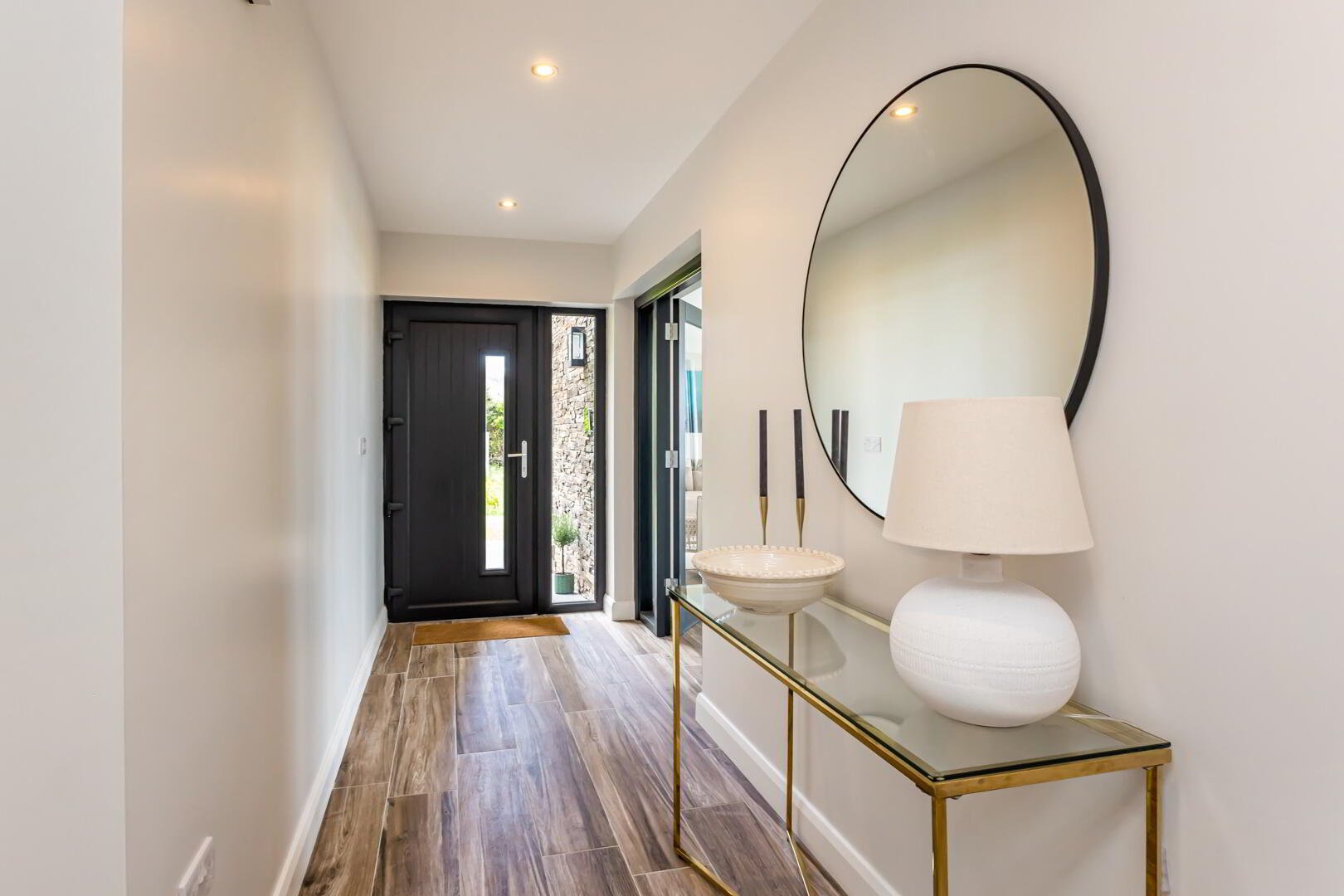 A modern entryway with a black front door, a glass panel, a gold and glass console table, and decor including a large round mirror, a table lamp, and a white bowl.
