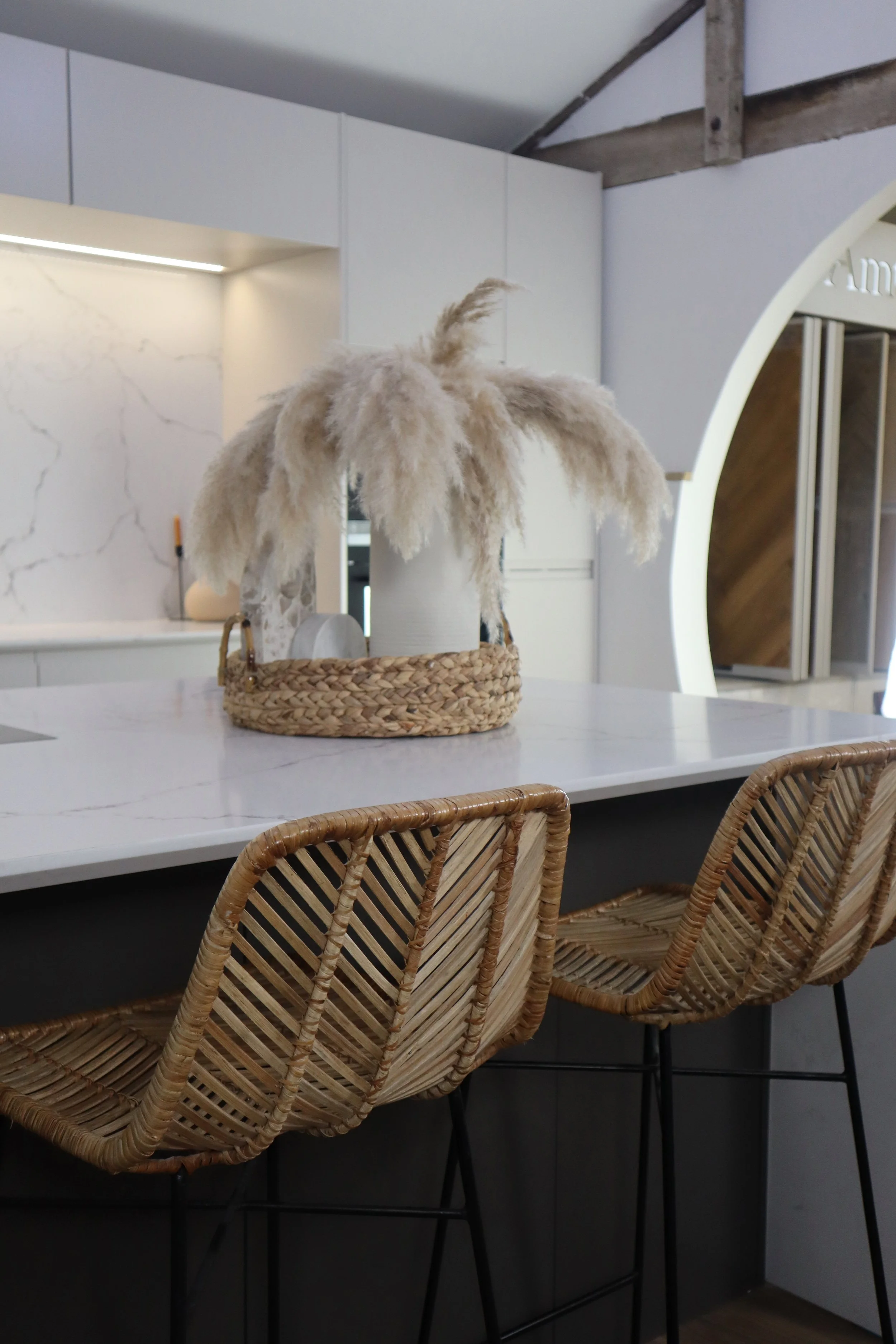 Kitchen island with a woven basket centerpiece holding a white vase with dried pampas grass, surrounded by two rattan bar stools.