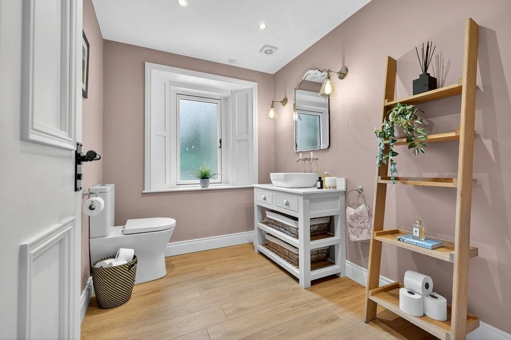 A bathroom with beige walls, wooden floor, white toilet, white vanity with a vessel sink, mirror, shelf, and a ladder-style shelving unit with decorative items and toiletries.