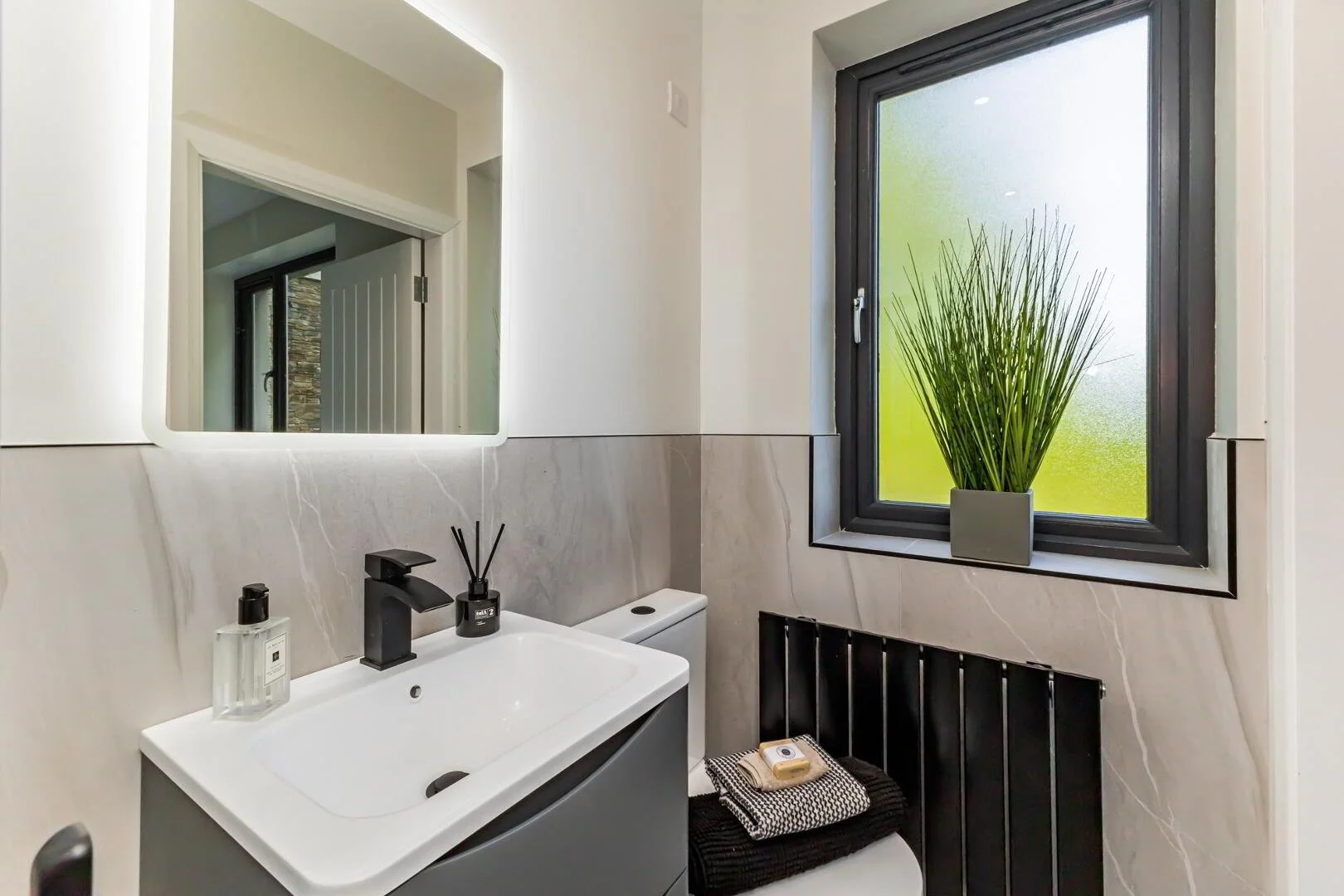 A modern bathroom with a white sink, black faucet, mirror, frosted window with black frame, black radiator, and a small green plant on the windowsill.
