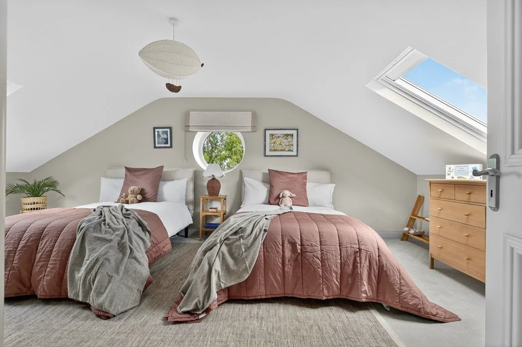 A cozy bedroom with two beds, brown bedding, plush toys, a small nightstand with a lamp, and a window showing trees outside. The room has a sloped ceiling with skylights and minimalist decor.