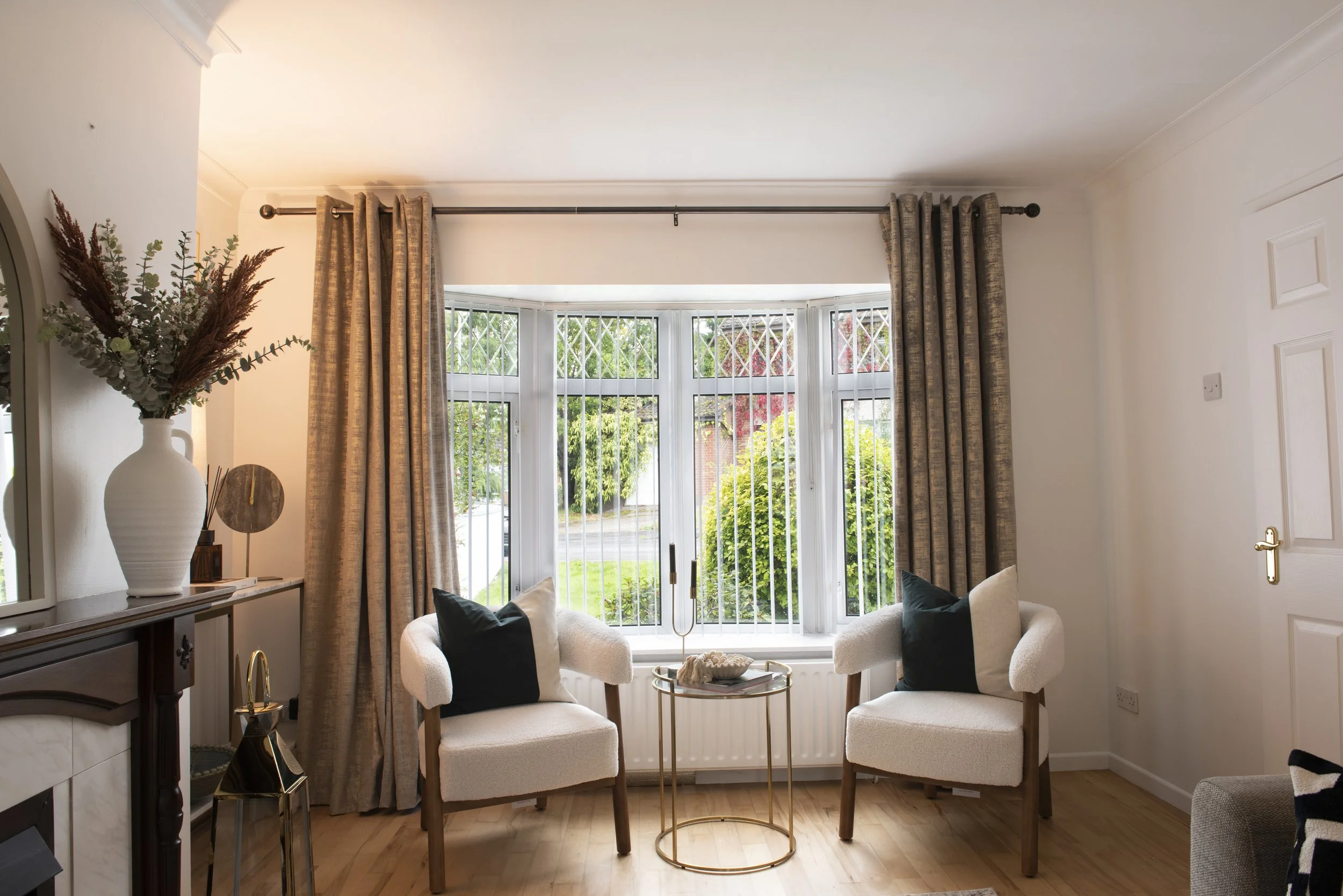 Interior living room with two cream-colored armchairs with black pillows, a small round gold table with decor, a bay window with beige curtains, decorative vase with foliage, and a window view of greenery outside.
