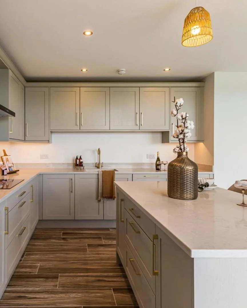 A modern kitchen with beige cabinets, a marble countertop island, a decorative vase with white flowers, and wooden flooring.