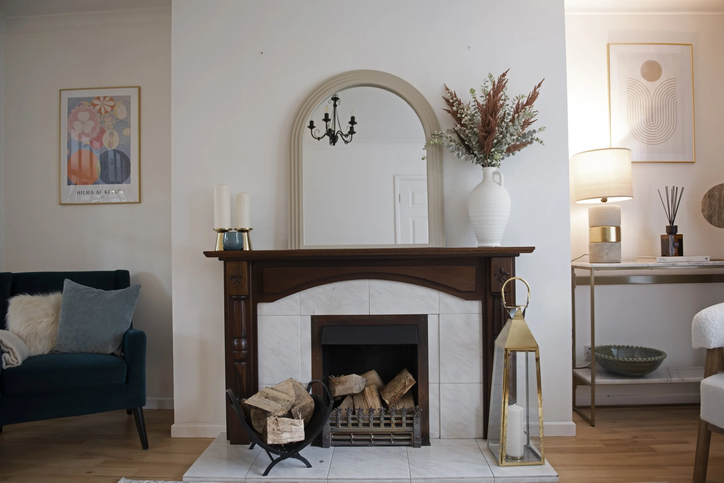 Living room with a wooden fireplace mantle, a large white vase with dried flowers, a mirror, artwork on the walls, and various lamps and decor.
