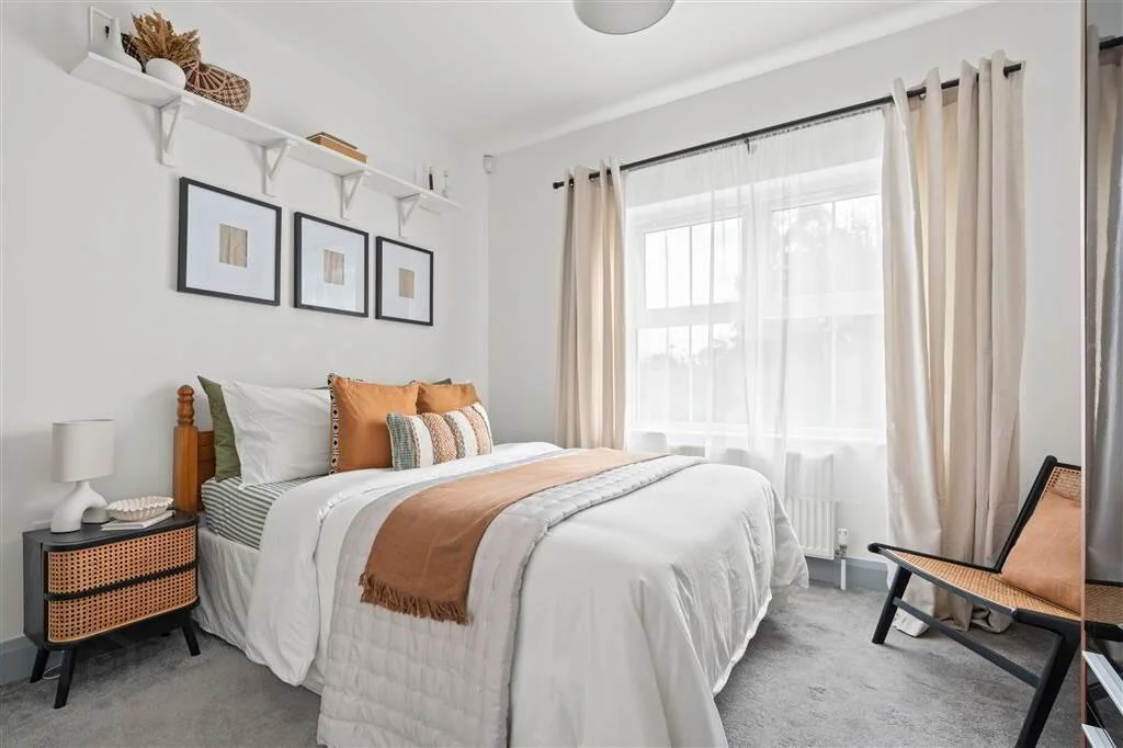A tidy bedroom with a large bed featuring white, beige, and rust-colored pillows and blanket. There are three framed abstract art pieces above the bed, a white windowsill with sheer curtains, and a small black nightstand with a white lamp. A wooden c