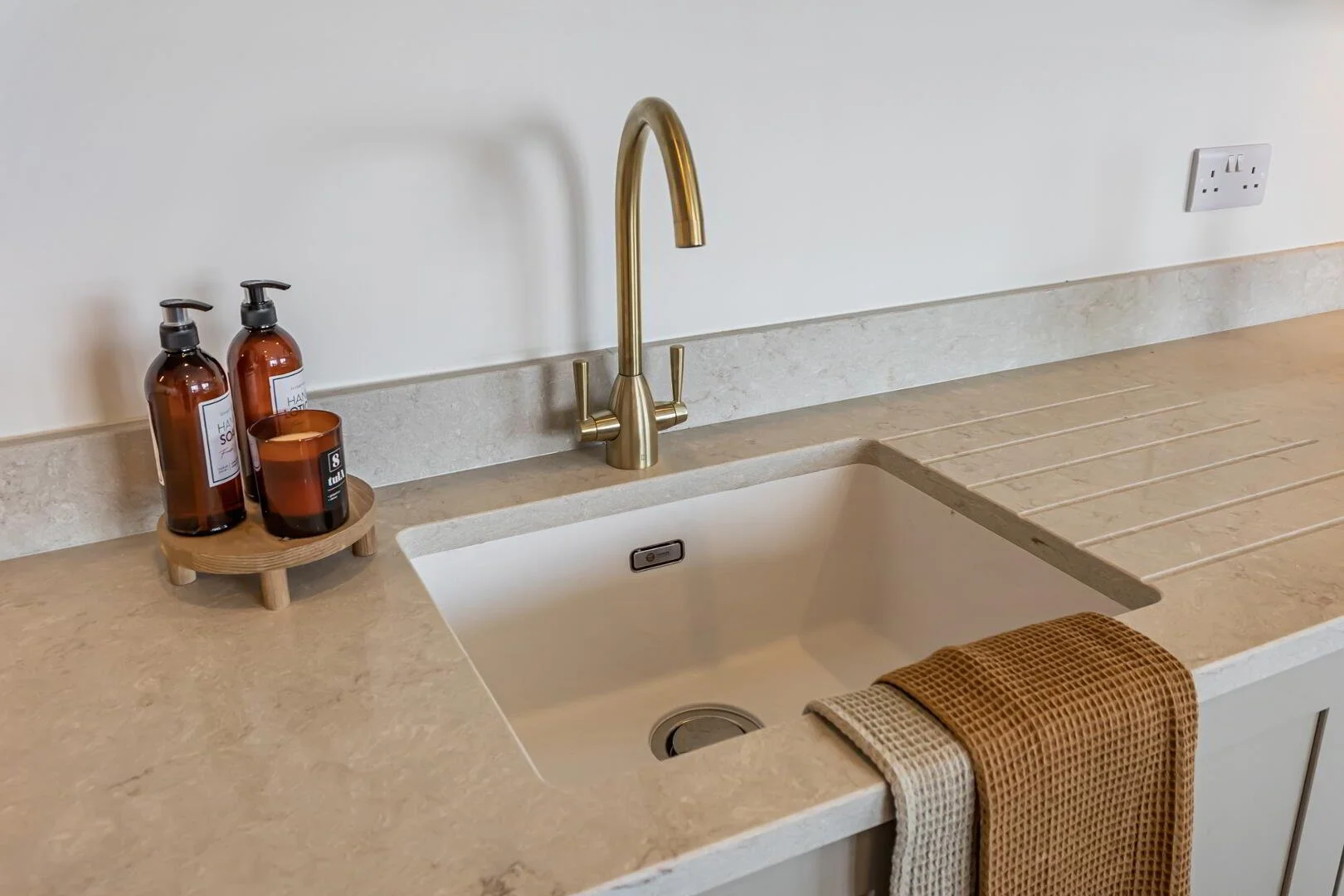 A kitchen sink with a brass faucet, beige countertop, a wooden tray with soap bottles, and a brown towel
