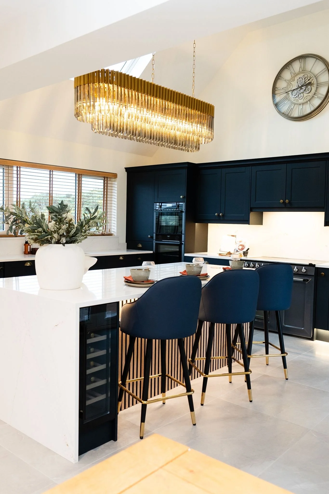 Modern kitchen with dark blue cabinets, white countertops, and a white island with three blue chairs. Bright chandelier hangs above, with a clock on the wall and a large window with blinds.