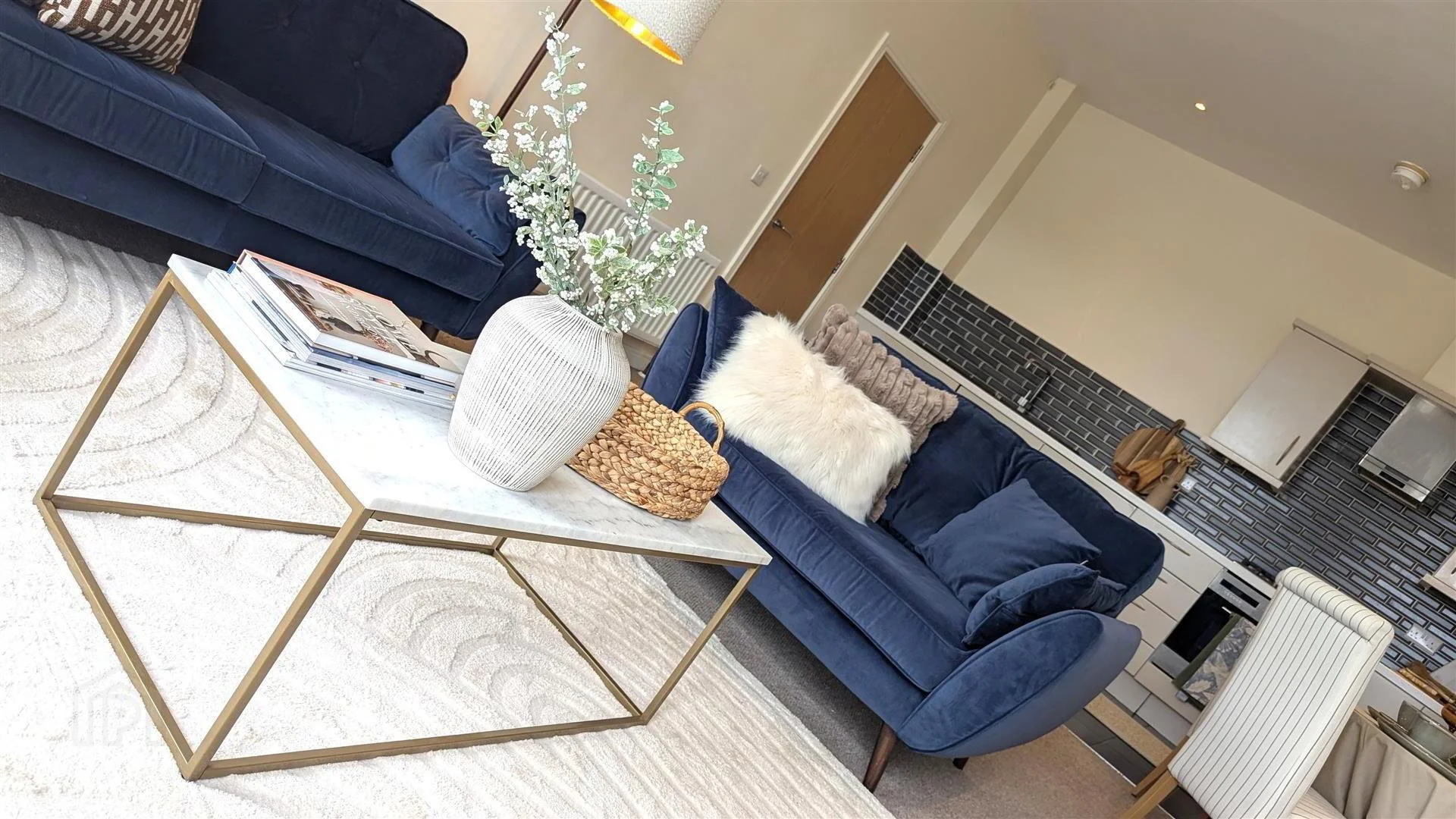 Living room with blue sofa, white coffee table with vase and magazines, kitchen in background with black backsplash and white cabinets, cream-colored wall, and a white dining chair.