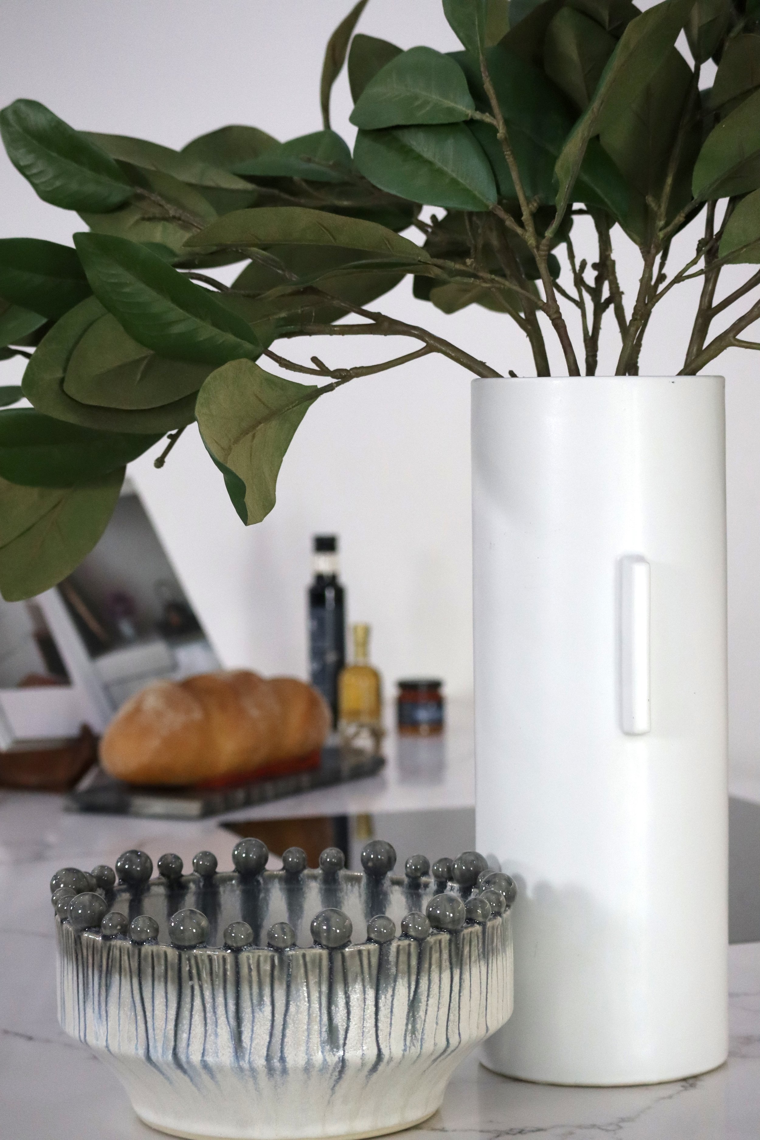 Vase with green leafy plant, decorative bowl with gray beads, and blurred background with bread and bottles.