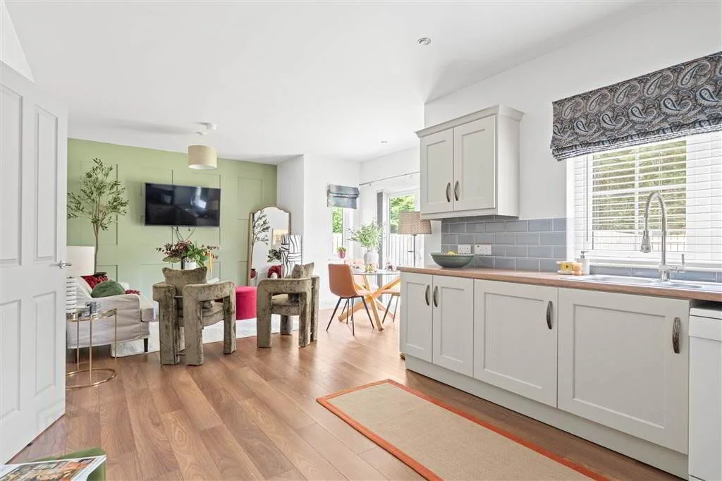 Open-concept living and kitchen area with hardwood floors, white cabinets, a window with blinds, a small rug, a dining table with orange chairs, a wall-mounted TV, and comfortable chairs with a small side table.