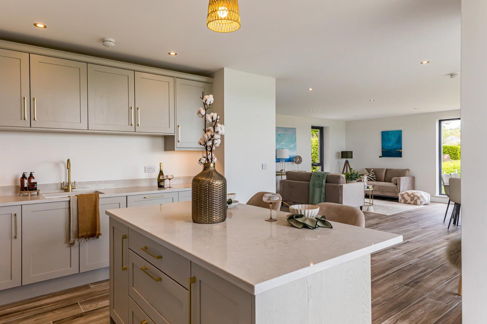 Open-concept living room and kitchen with light-colored cabinetry, a large kitchen island with a vase of cotton branches, beige sofas, and artwork on the walls
