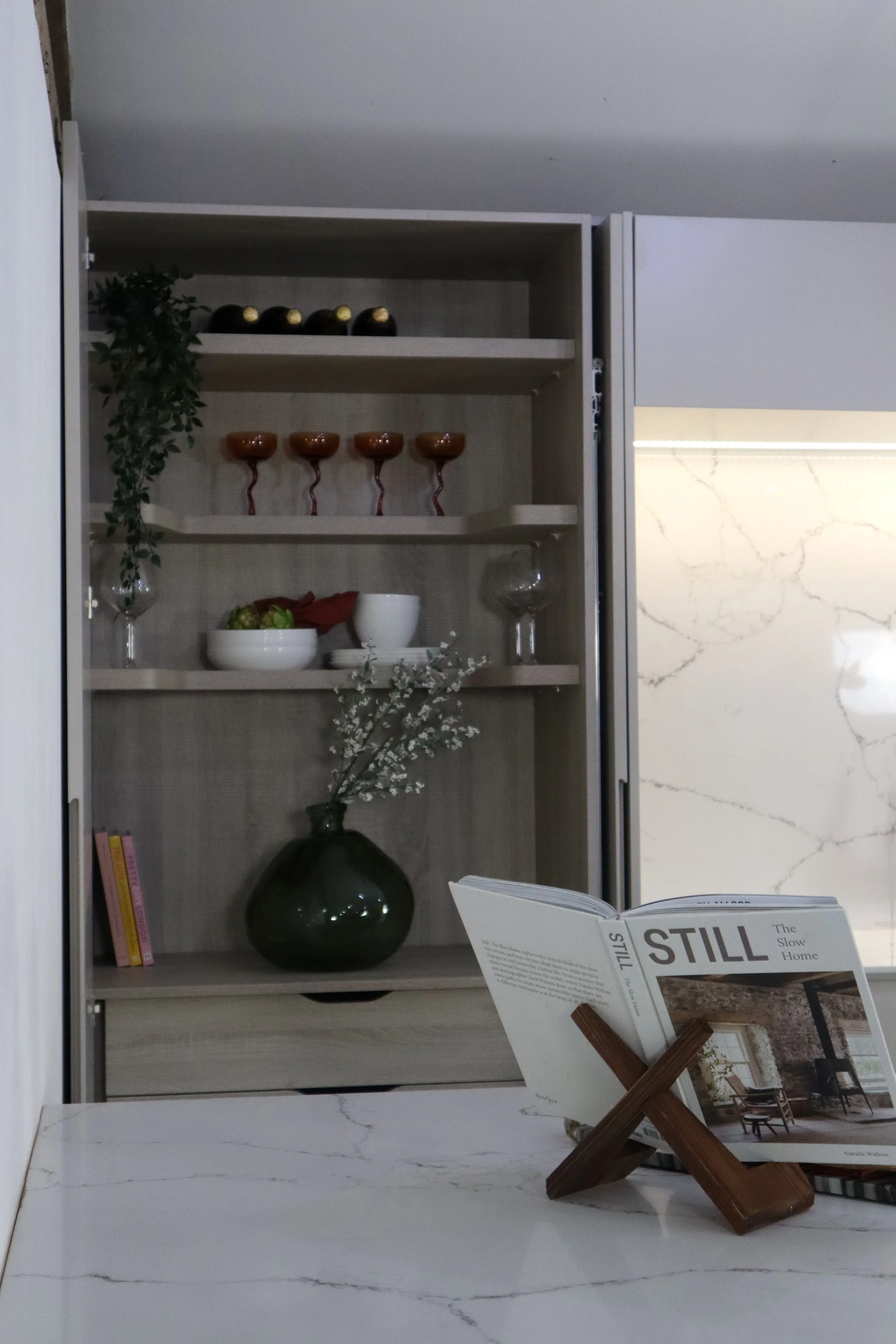 Open wooden cabinet displaying decorative items including black and gold spheres, orange chalices, glassware, and a white bowl with green fruit. In the foreground, a white marble countertop holds an open magazine titled 'STILL', propped up by a small wooden stand, with a green vase and white flowers behind it.