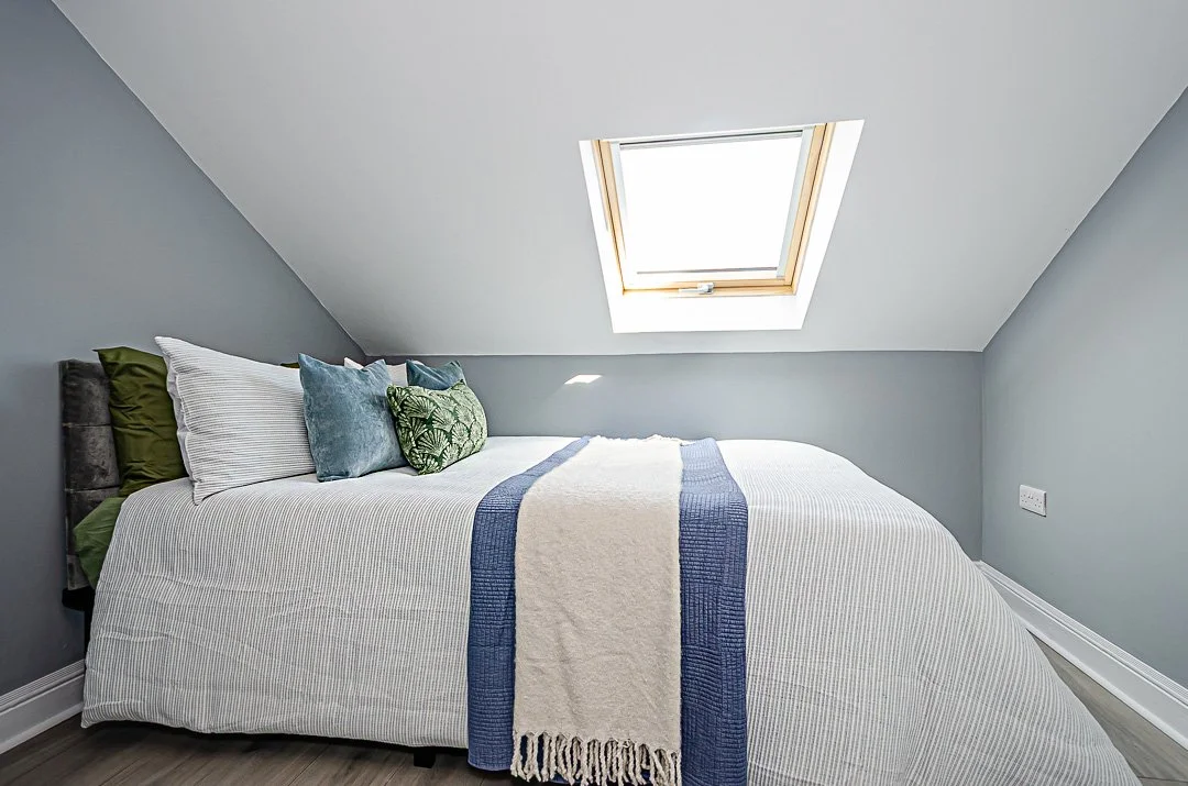 A cozy bedroom with a large bed, gray walls, and a skylight window in the ceiling. The bed has striped bedding and several pillows, including green, white, blue, and patterned ones. There is a white and blue throw blanket on the bed.
