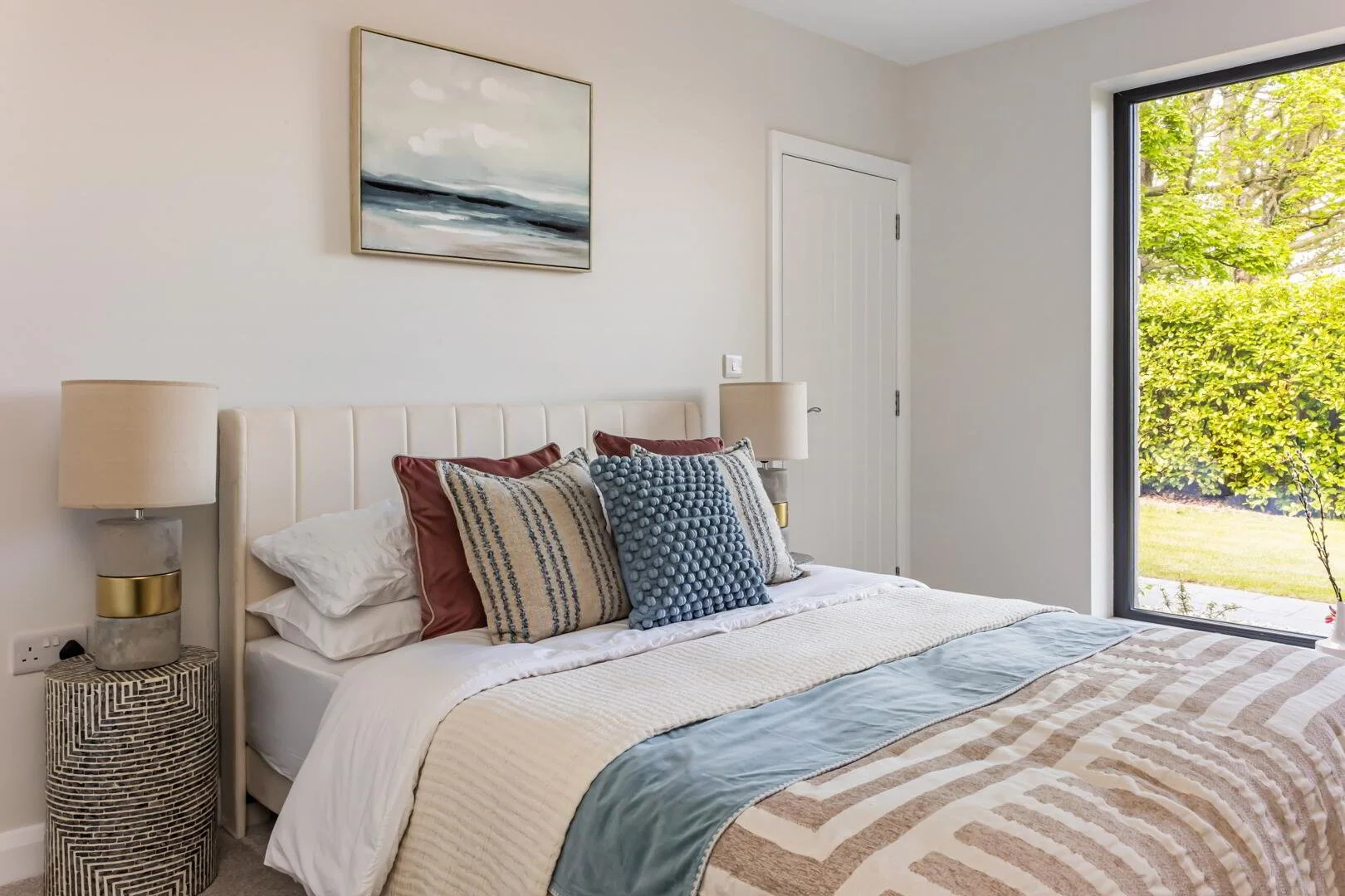 A bedroom with a bed, multiple pillows, bedside lamps, a window showing outside greenery, a door, and a landscape painting on the wall.