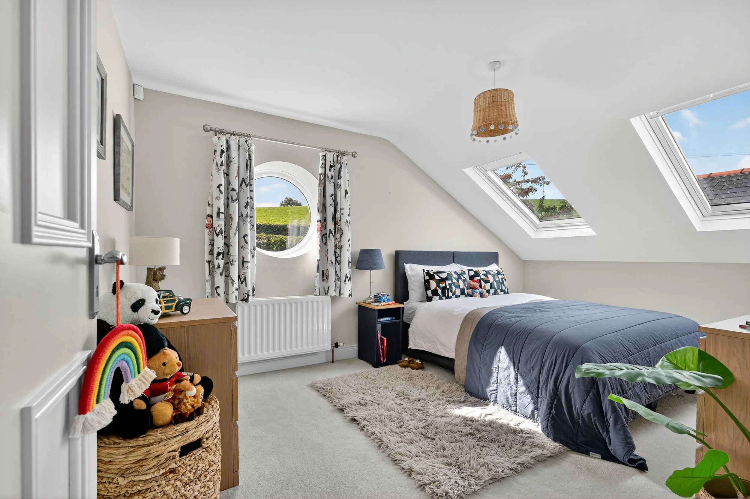 A cozy attic bedroom with a bed, colorful pillows, a navy blue comforter, a fluffy rug, a window with patterned curtains, skylights, and a side table with a blue lamp.