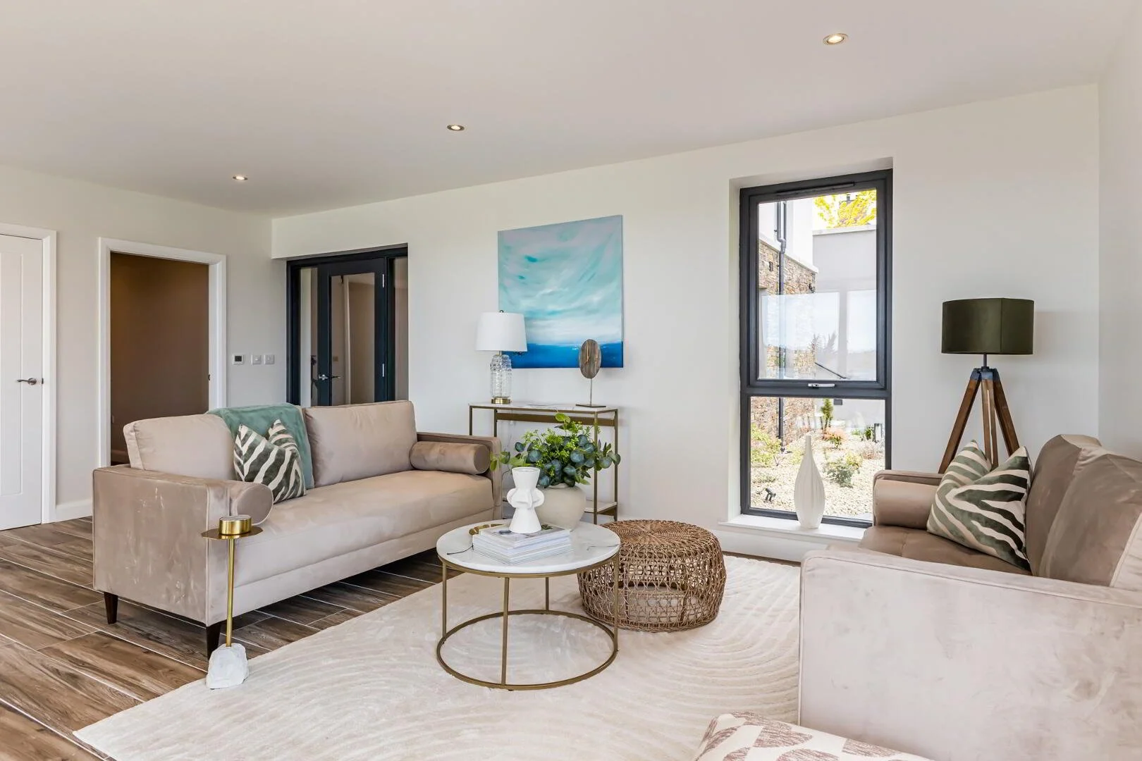 Living room with beige sofas, a white rug, a round coffee table with decorative items, and a large window showing an outdoor garden.
