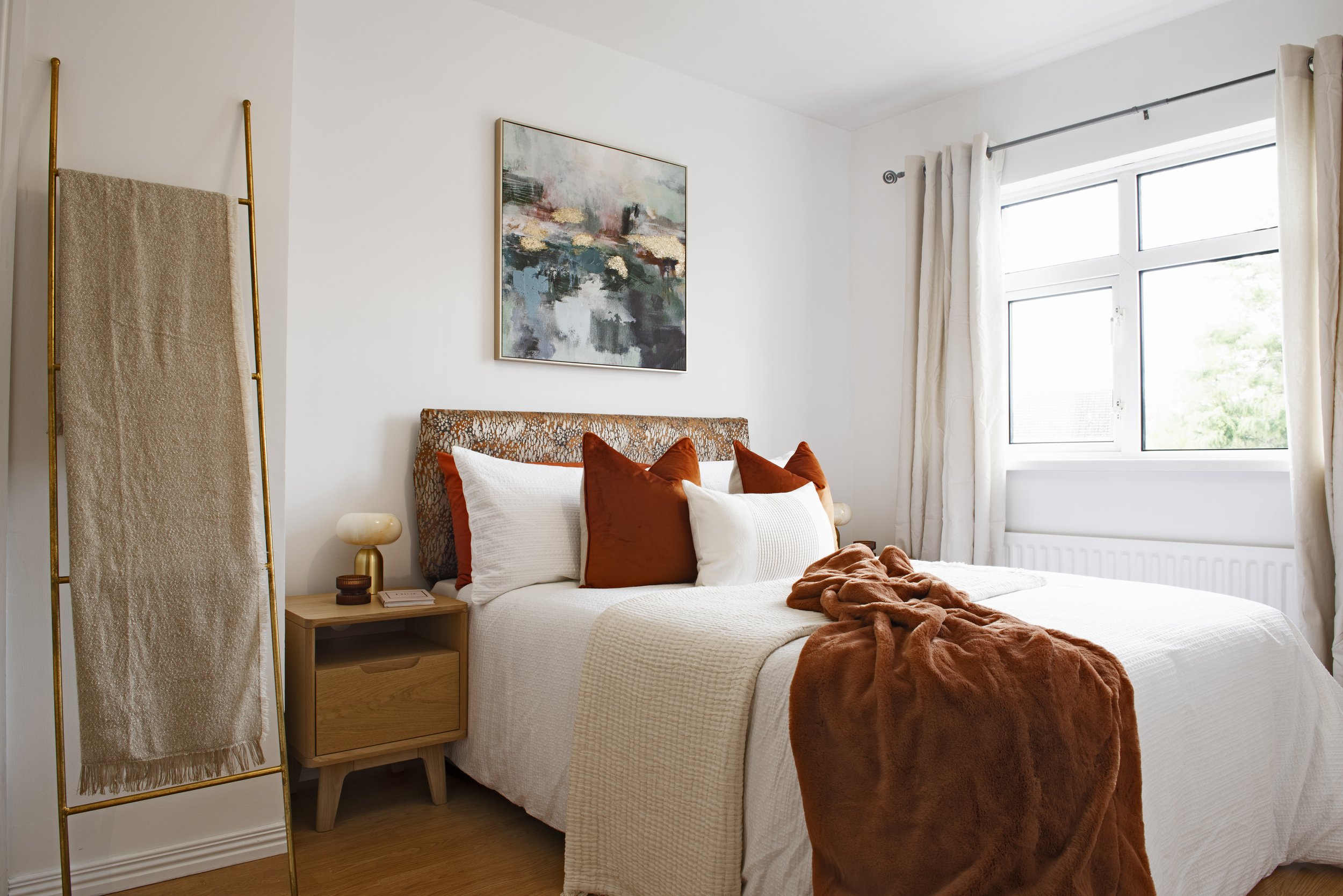 A bedroom with a white bed covered in a beige blanket, decorated with white and rust-colored pillows. Next to the bed is a wooden nightstand with a lamp and a book. A window with white curtains lets in natural light. A framed abstract painting hangs 