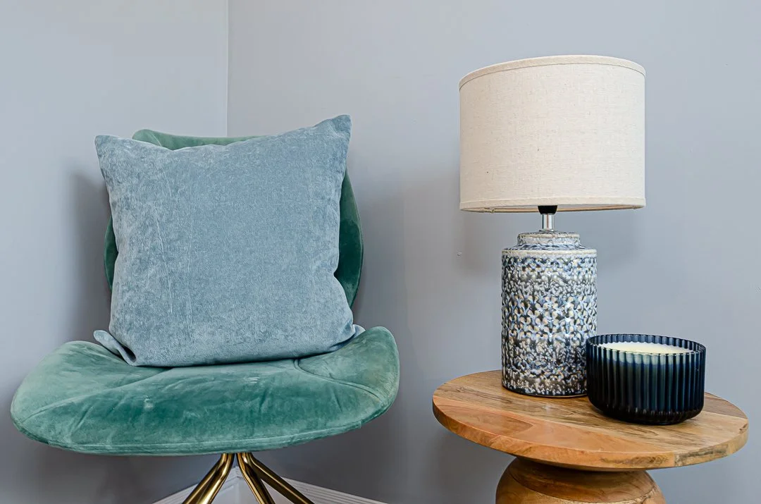 A green velvet chair with a large blue pillow, a wooden side table with a decorative lamp and a black bowl on top, against a light blue wall.