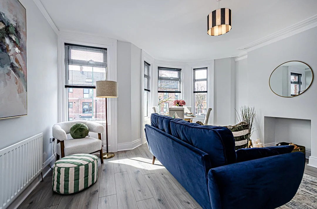 Bright living room with bay windows, white walls, dark blue sofa, white armchair with green pillow, striped ottoman, round mirror, and decorative plants.