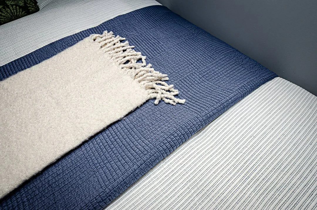 A bed with a textured blue bedspread, a white and blue striped sheet, and a cream-colored blanket with fringes.