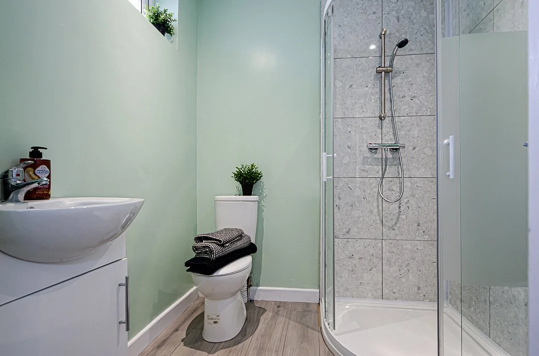 Small bathroom with light green walls, a white sink with a soap dispenser, a white toilet with a black and white towel on top, a potted plant on the toilet tank, and a shower with a clear glass door and gray tiled walls.