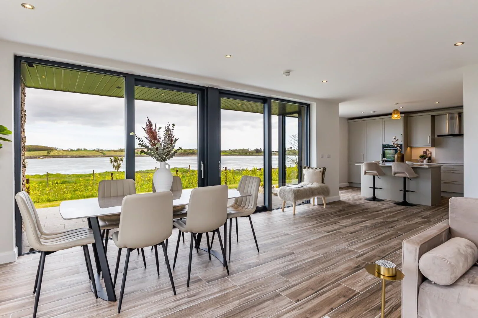 Modern living room with large glass sliding doors showing a river view, dining table with chairs, a bench with a fur throw, and a kitchen area with bar stools.