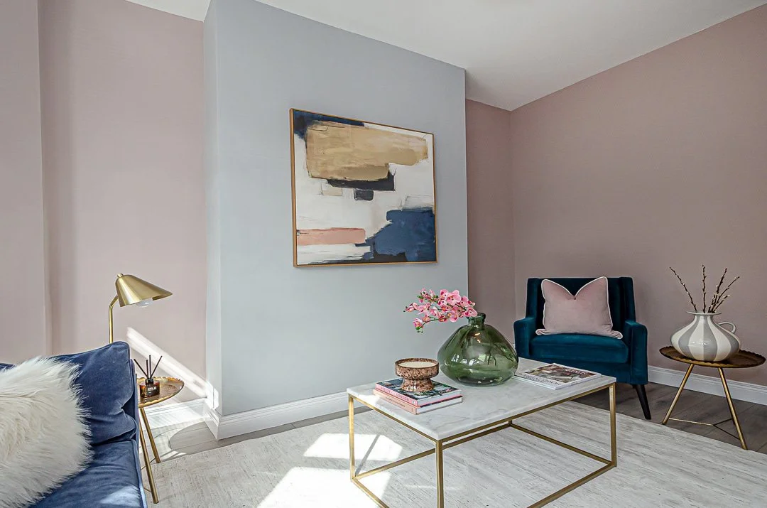 Living room with modern decor, blue and pink walls, a blue armchair with a pink pillow, a marble-top coffee table with pink flowers and books, a gold-side table with a lamp, and a white vase with branches.