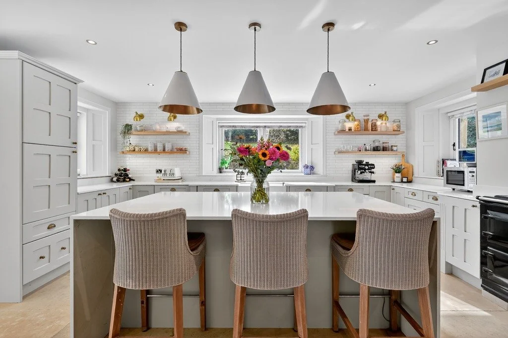 Bright white kitchen with a large island, hanging pendant lights, and open shelves with decorative items, flowers in the center, window view outside, and kitchen appliances.