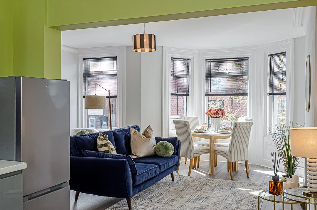 Bright living and dining area with large bay windows, a round dining table with four striped chairs, a navy blue sofa with various cushions, a small side table with a lamp and decorative items, and a modern pendant light.