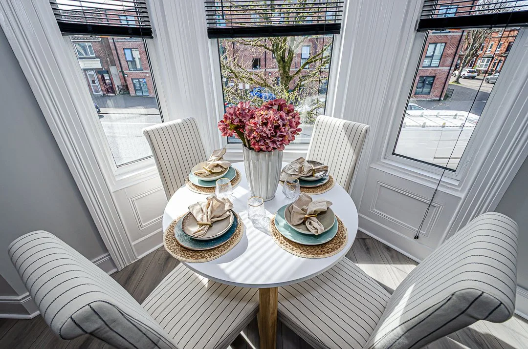 A small round dining table set for four with beige napkins, glasses, and dinnerware, featuring a large vase with pink hydrangeas. The table is positioned in a sunlit corner with large windows showing an urban street view.
