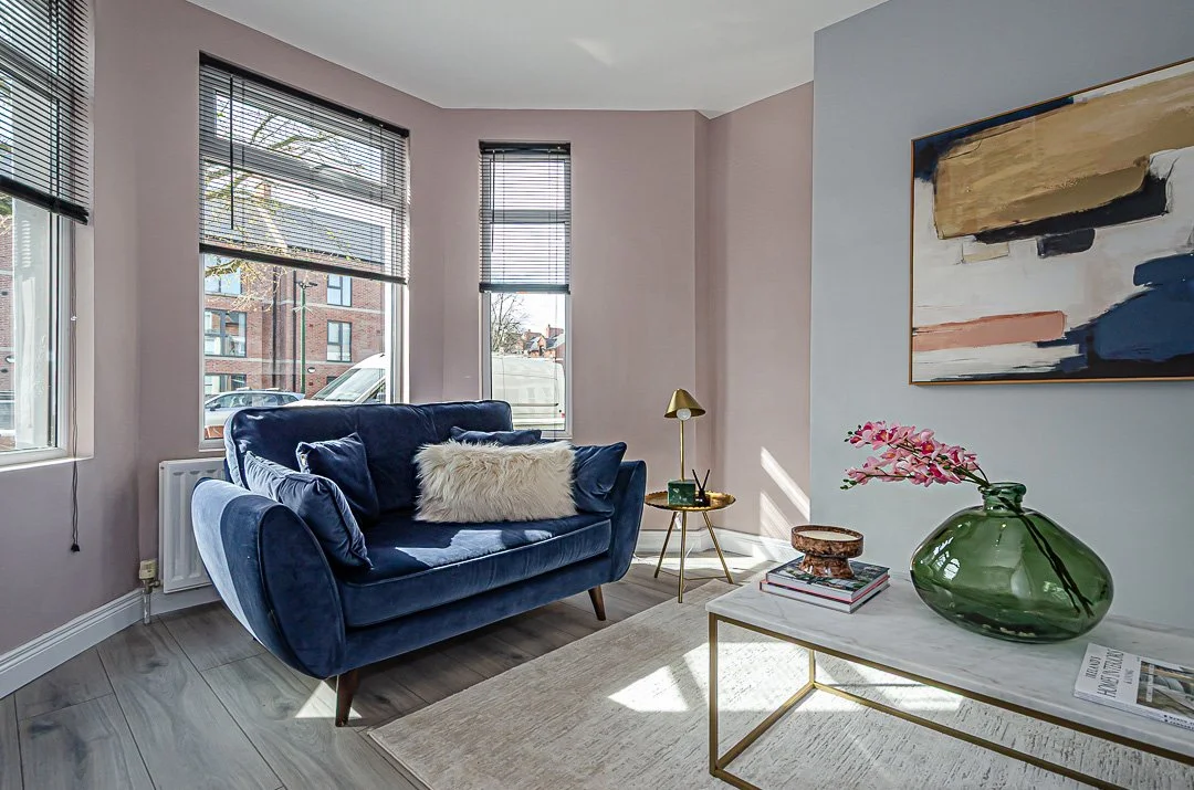 Living room with large windows, navy blue velvet sofa, fur and velvet pillows, modern side table with gold lamp, abstract painting on wall, vase with pink flowers on a white coffee table, wooden floors, and beige area rug.