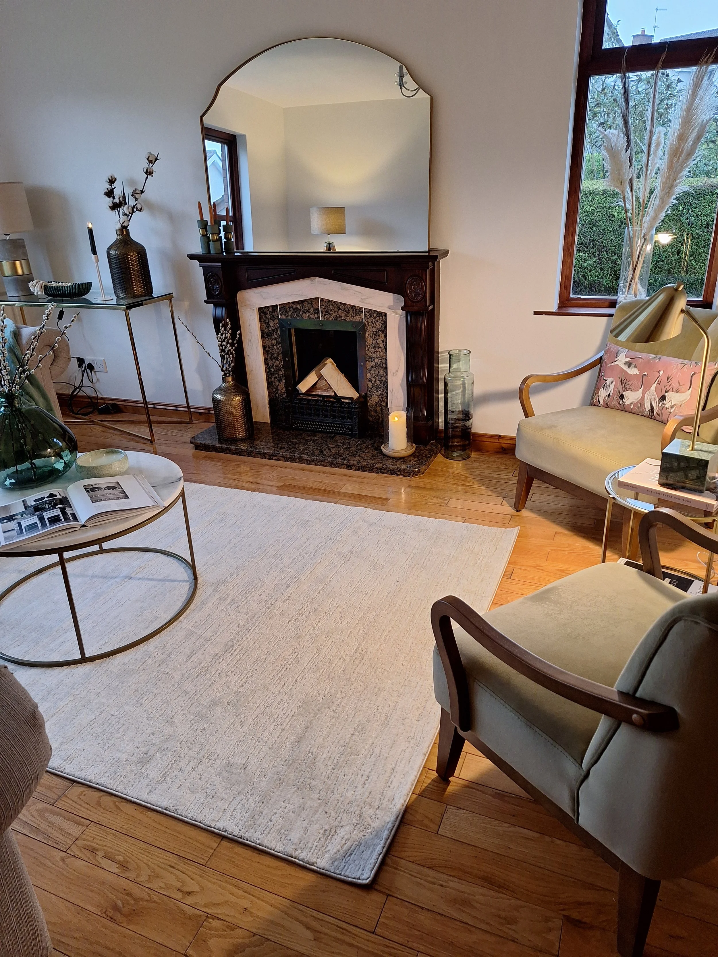A cozy living room with a fireplace, a white rug, and various chairs and tables. There is a large mirror above the fireplace, and decorative items such as vases, candles, and a lamp are placed around the room. The room has wooden flooring and a windo